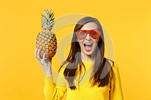 Excited young woman in funny glasses keeping mouth open, hold fresh ripe pineapple fruit isolated on yellow orange wall