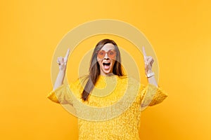 Excited young woman in fur sweater and heart orange glasses pointing index fingers up on copy space isolated on bright