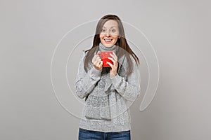 Excited young woman in gray sweater, scarf holding red cup of coffee or tea isolated on grey wall background. Healthy