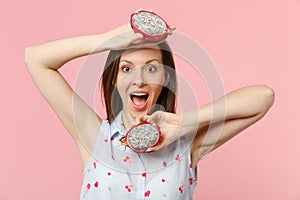 Excited young woman keeping mouth open, holding halfs of fresh ripe pitahaya, dragon fruit isolated on pink pastel wall