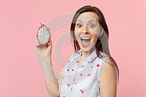 Excited young woman keeping mouth wide open holding half of fresh ripe pitahaya dragon fruit isolated on pink pastel