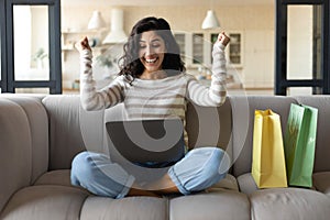 Excited young woman with laptop and shopping bags making YES gesture, buying things online from home