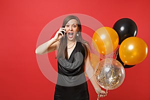 Excited young woman in little black dress holding air balloons, talking on mobile phone, screaming isolated on bright