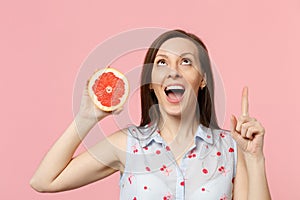 Excited young woman looking, pointing index finger up holding half of fresh ripe grapefruit isolated on pink pastel wall