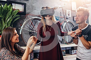 Excited young woman playing interactive videogame wearing virtual reality headset with friends holding her hands