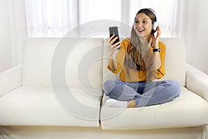 Excited young woman sitting with legs crossed on couch watching music video on smartphone at home. Copy space