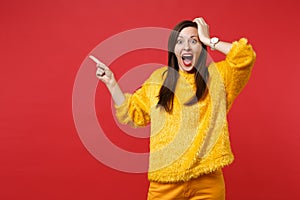 Excited young woman in yellow fur sweater screaming, pointing index finger aside, putting hand on head isolated on red