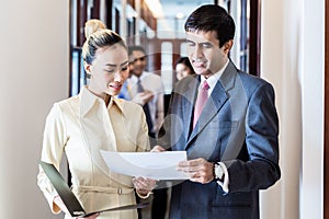 Executive and secretary standing in office hallway discussing papers