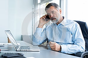 Executive senior businessman using his mobile phone and talking wih somebody while working laptop in the office.