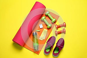 Exercise mat, dumbbells, bottle of water, wireless earphones, fitness elastic band and shoes on yellow background, flat lay
