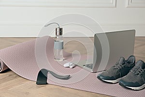 Exercise mat, laptop, bottle of water, wireless earphones, fitness elastic band and shoes on wooden floor indoors