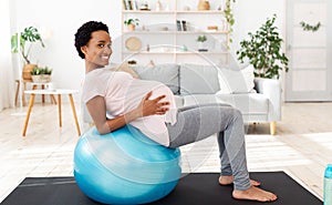 Exercising during pregnancy concept. Positive black future mother working out on fitness ball at home