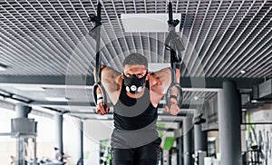 Exercising on rings. Young sportive strong man in black wear and mask have workout day in gym