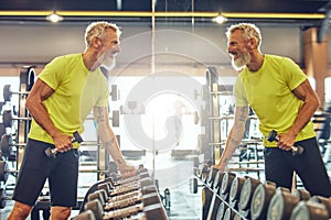 Exercising with weights. Side view of a handsome mature man in sportswear taking a dumbbells, looking in a big mirror