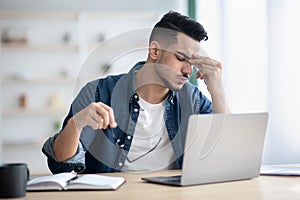 Exhausted arab guy sitting at workdesk, removing glasses