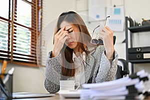 Exhausted Asian businesswoman at her desk, taking off her eyeglasses, touching nose bridge