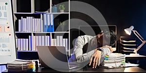 Exhausted businessman sleeping at his desk and working overtime late at night, he is surrounded by piles of paperwork