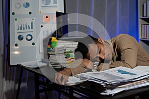 Exhausted businessman sleeping at his desk and working overtime late at night, he is surrounded by piles of paperwork