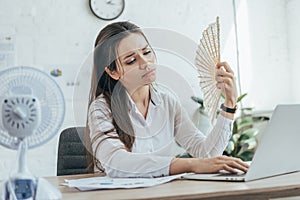 exhausted businesswoman using laptop while conditioning air with electric fan and hand fan