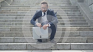 Exhausted Caucasian businessman walking down the stairs and sitting down. Wide shot portrait of tired young man in