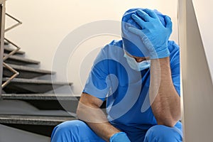 Exhausted doctor sitting on stairs indoors. Stress of health care workers during COVID-19 pandemic