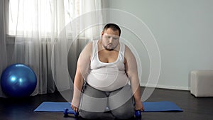 Exhausted fat man sitting on floor with dumbbells, tiring workout program, sport