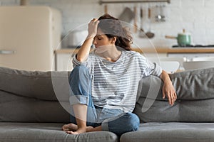 Exhausted frustrated young woman touching forehead, sitting on couch alone