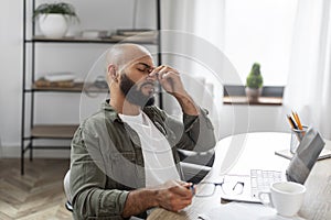 Exhausted latin male employee working on laptop at office, sitting at workdesk and touching nose bridge, feeling tired