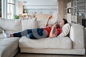 Exhausted man with difficult breath lying on sofa, waving with hand fan in need of cool, fresh air