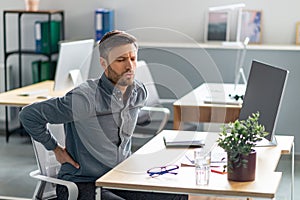 Exhausted mature businessman having acute back pain, massaging aching muscles while sitting at desk in office