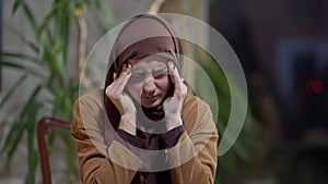 Exhausted Muslim woman closing laptop holding head in hands and stretching sitting in home office in the evening. Medium