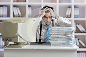 Exhausted overworked man is looking on pile of documents on desk. Paperwork concept.