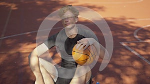 Exhausted perspiring redhead sportsman sitting with ball on outdoor court and looking up at sunlight. Portrait of tired