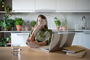 Exhausted sleepy woman having work laptop break, holding smartphone, reading news, procrastinating