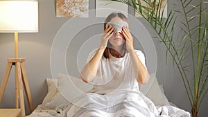 Exhausted sleepy young woman sitting in bed taking off blindfold feeling drowsy after wake up too early in morning, sleepless