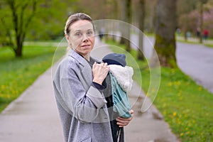 Exhausted and stressed late-term mother in her 40s holding her newborn baby in a baby carrier and looking annoyed to the camera