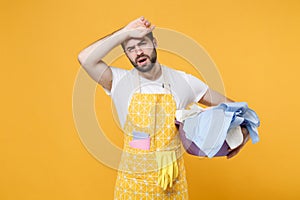 Exhausted tired young man househusband in apron hold basket with clean clothes while doing housework isolated on yellow