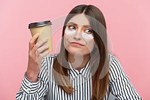Exhausted unmotivated woman in striped shirt drinking hot beverage from paper cup, sitting with eye patches under eyes, coffee