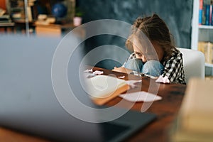 Exhausted upset primary child school girl sitting alone hugging knees in front of desk with difficult homework.