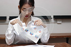 Exhausted upset young Asian business female tearing up charts or paperwork