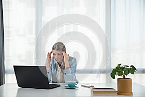 Exhausted upset young business woman sitting at table with laptop, leaning head on hand, suffering from headache, feeling depresse