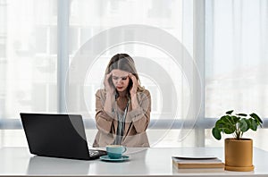 Exhausted upset young business woman sitting at table with laptop, leaning head on hand, suffering from headache, feeling depresse