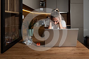 Exhausted upset young student girl sitting at table with laptop, leaning head on hand, suffering from headache, feeling depressed