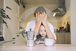 Exhausted young woman with baby is sitting with coffee in kitchen. Modern tired mom and little child after sleepless night. Life photo