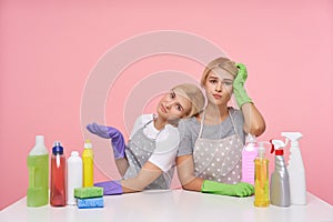 Exhausted young attractive blonde cleaning ladies wearing working clothes and rubber gloves while sitting over pink background,