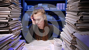 Exhausted young business woman office employee at the desk working late at night overloaded with paperwork. Female works