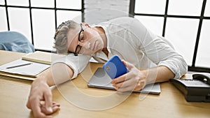 Exhausted young caucasian businessman, smartphone in hand, loses battle with stressâ€“ nobly crashes asleep on office table,