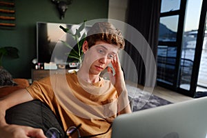 Exhausted young caucasian man holding eyeglasses by laptop on sofa in living room at home