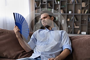 Exhausted young male recline on sofa hold fan wave himself