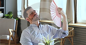 Exhausted young man waving fan suffering from summer heat indoors